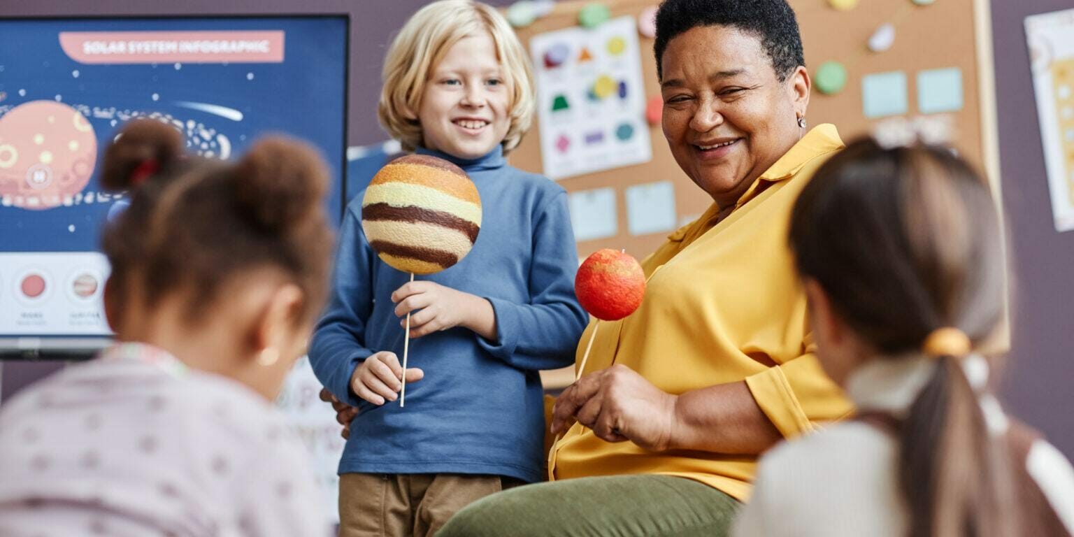 A smiling childcare teacher engaging with young children in a hands-on space-themed learning activity. A boy holds a model of Jupiter while the teacher holds a model of Mars, fostering an interactive and joyful educational experience.
