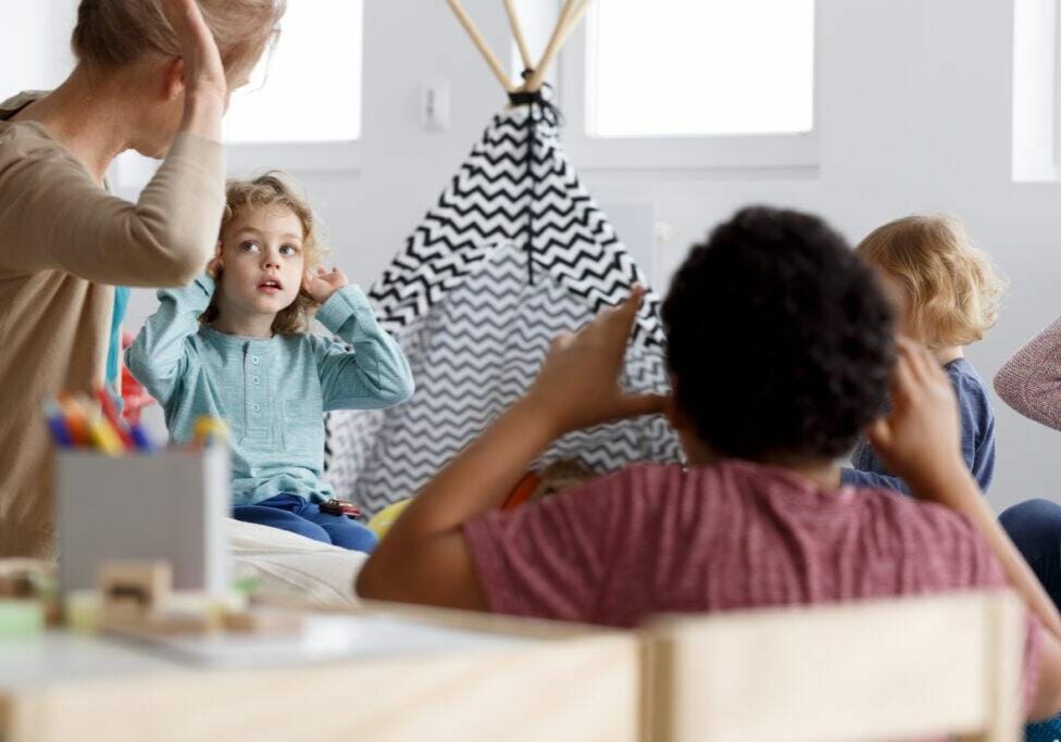 A caring childcare teacher engaging a group of young children in an interactive learning activity in a cozy, well-lit classroom. A young girl attentively follows along, mirroring the teacher’s gestures, while other children participate. The setting includes a small indoor play tent and various educational materials, fostering a warm and supportive environment.