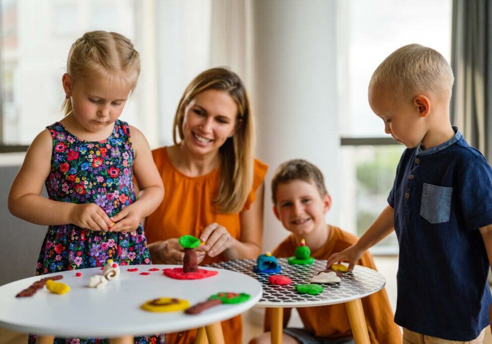 smiling childcare provider engaging with young children in a creative play activity using colorful modeling clay. The children are focused on shaping and designing objects, fostering imagination and hands-on learning in a warm, nurturing environment.