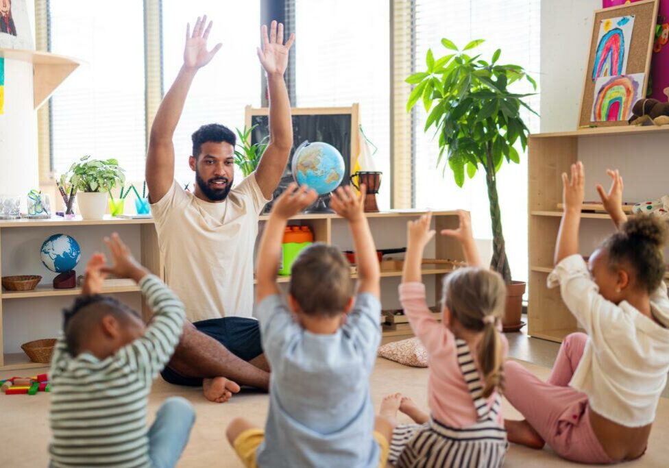 A caring male teacher leading a fun and interactive learning activity with a group of young children in a bright and engaging classroom. The children sit on the floor with arms raised, participating enthusiastically. The setting includes educational materials, plants, and children’s artwork, creating a warm and supportive learning environment.